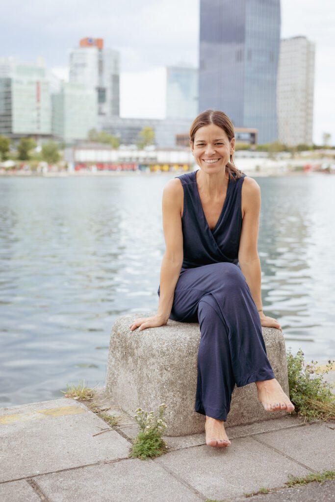 katrin_kindul_freudvoll_beziehungen_gestalten Katrin sitzt freudvoll, lachend auf einem quadratischen Stein. Im Hintergrund die Donau und die Wiener Wolkenkratzer.